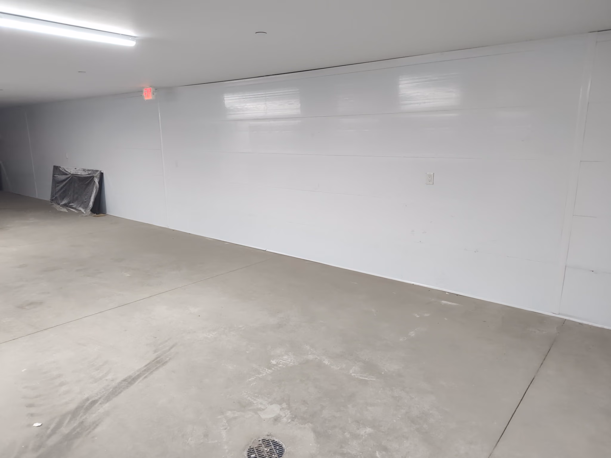 Empty white indoor room with concrete floor, a floor drain, and a covered object in the corner.