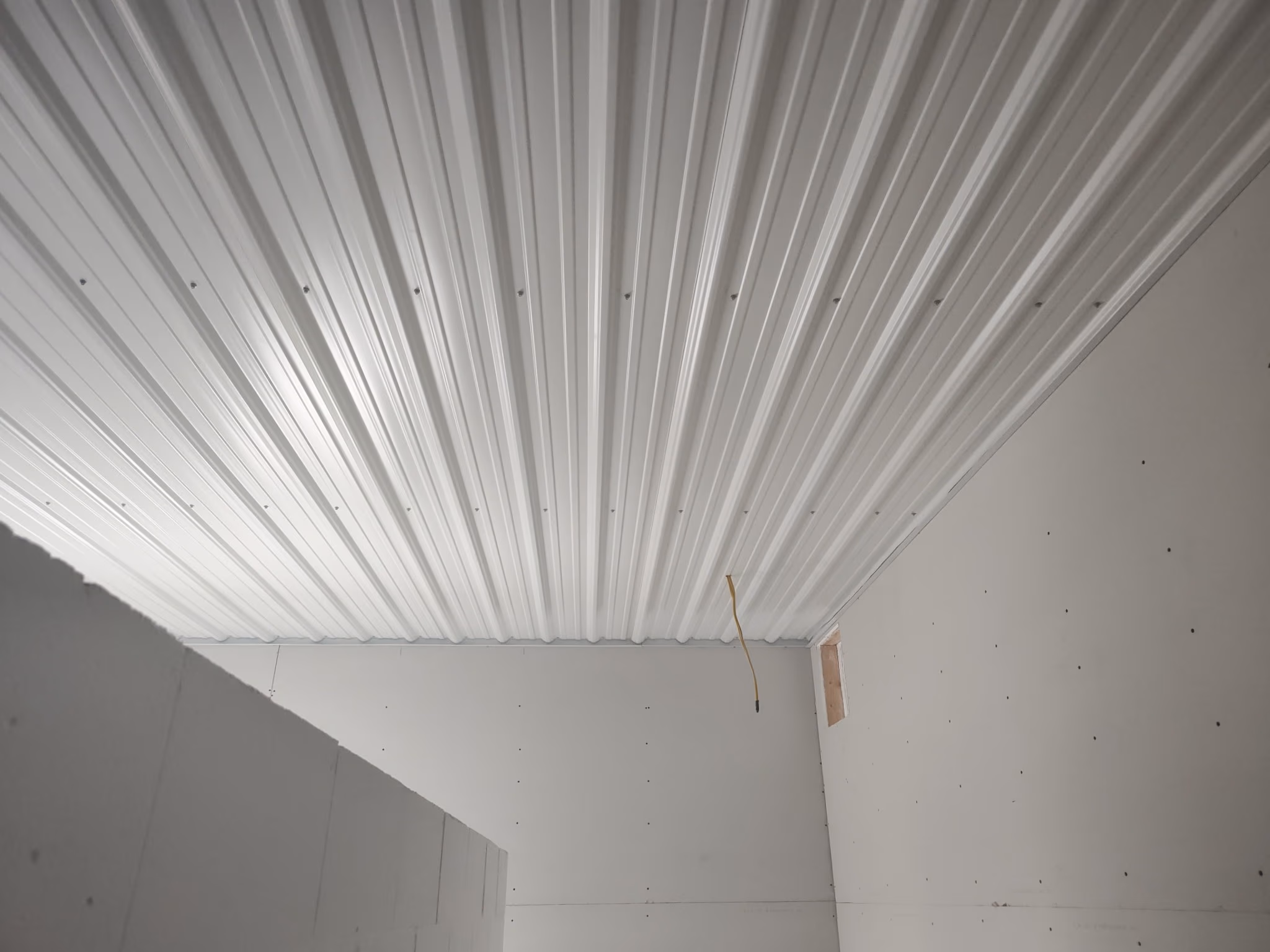 Indoor room under construction with white corrugated metal ceiling and unfinished drywall walls.